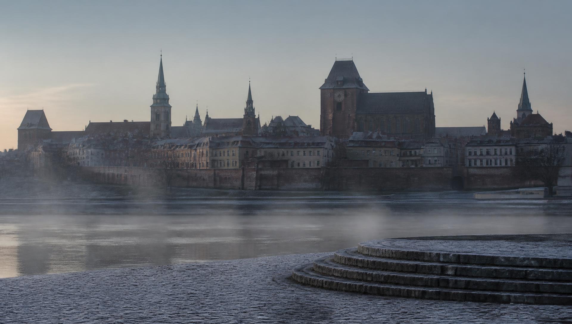 Dawn over the Gothic skyline of Toruń's medieval Old Town, mist rising from the Vistula river.